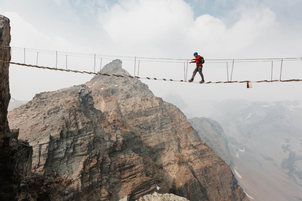 A man crosses a long suspension bridge between two peaks on the Mt. Nimbus via ferrata at CMH Bobbie Burns.