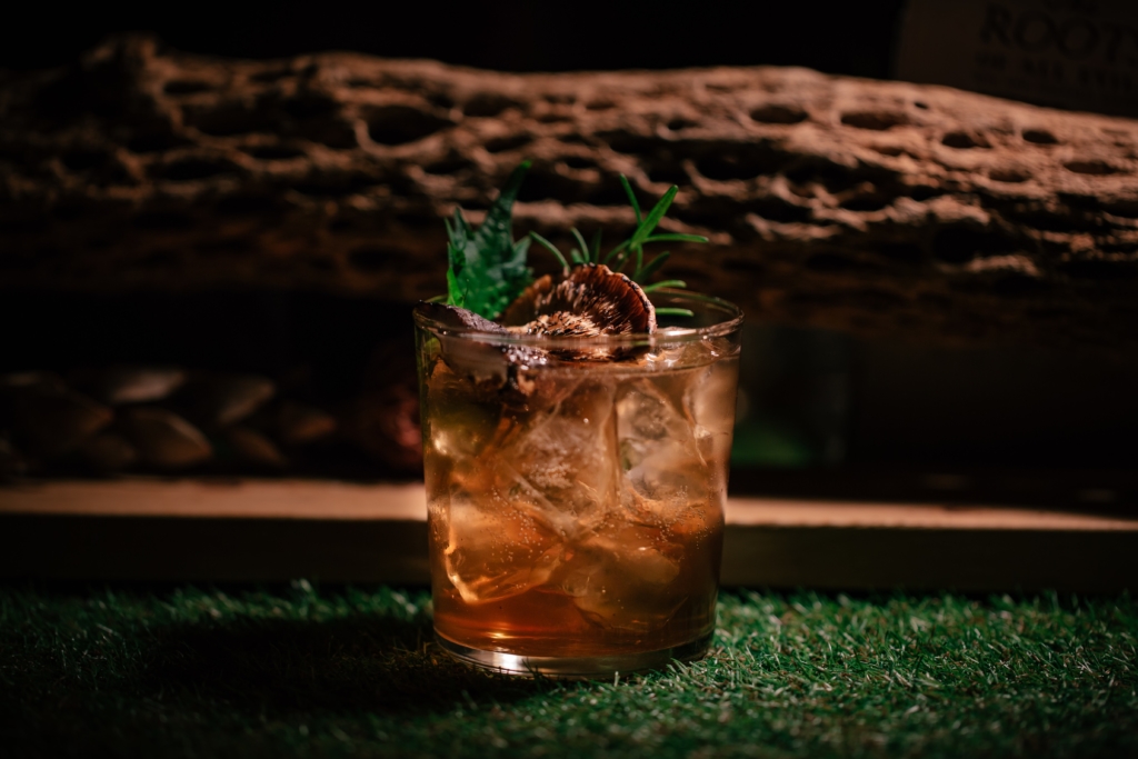 A deep brown cocktail with lots of ice sits on grass in front of a log