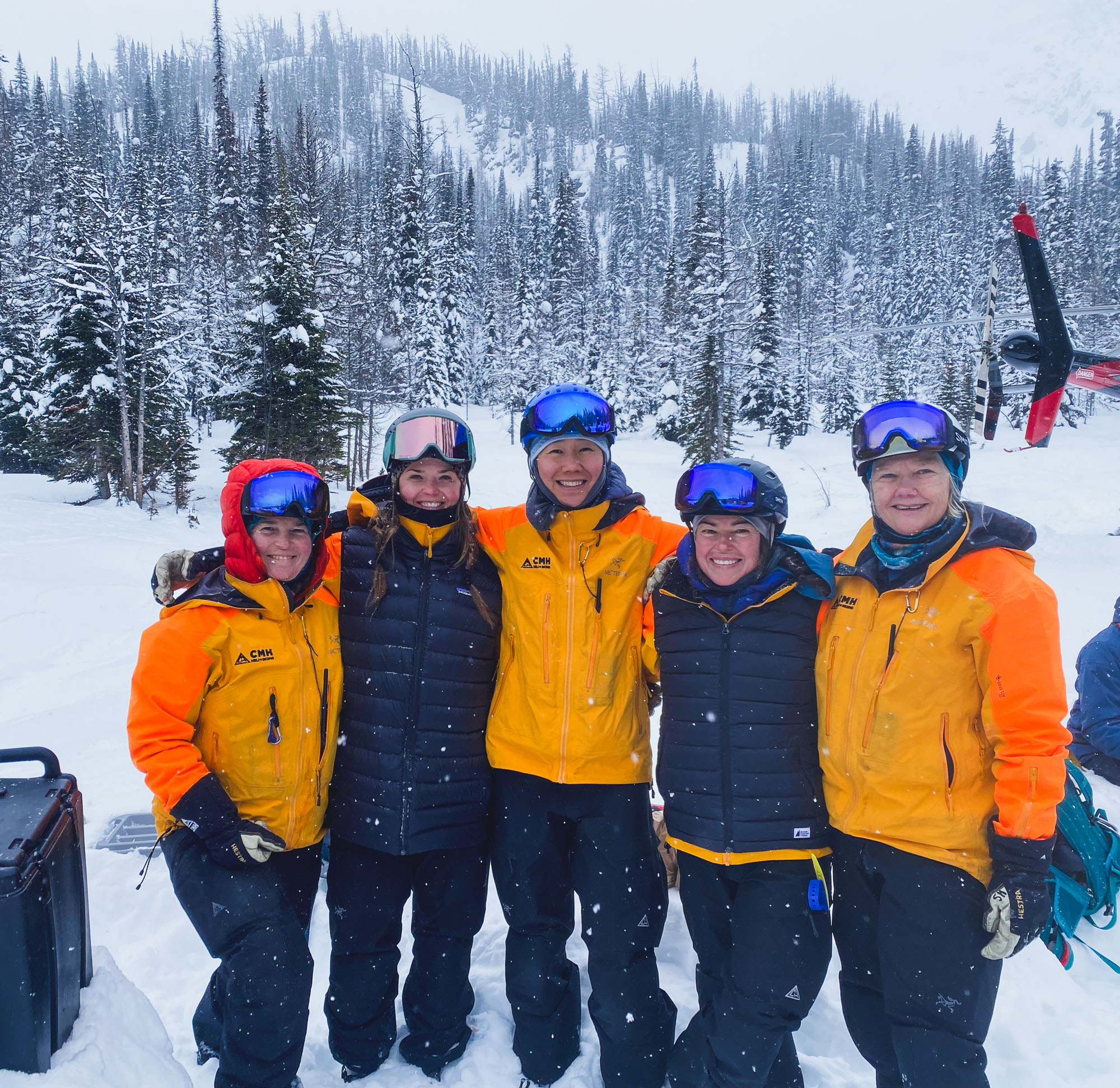 Five female mountain guides wearing bright orange jackets pose for a group photo after a day of guiding heli-skiing at CMH Bugaboos in British Columbia, Canada. There's forest, snow and part of a helicopter visible in the background.