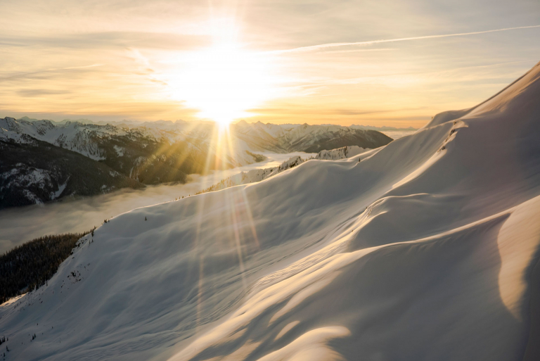 The sun setting over a snowy mountain vista, seen from a helicopter.