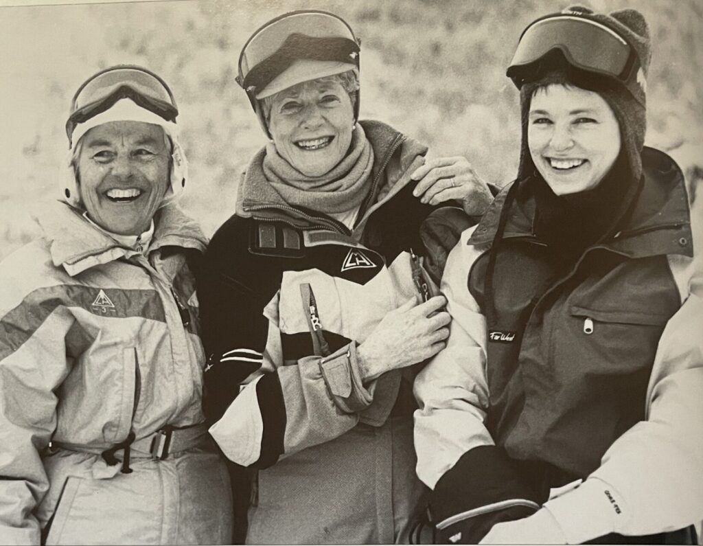 An aged, sepia photograph shows three smiling middle-aged women. They are wearing ski goggles and Million Foot ski suits from CMH Heli-Skiing. 