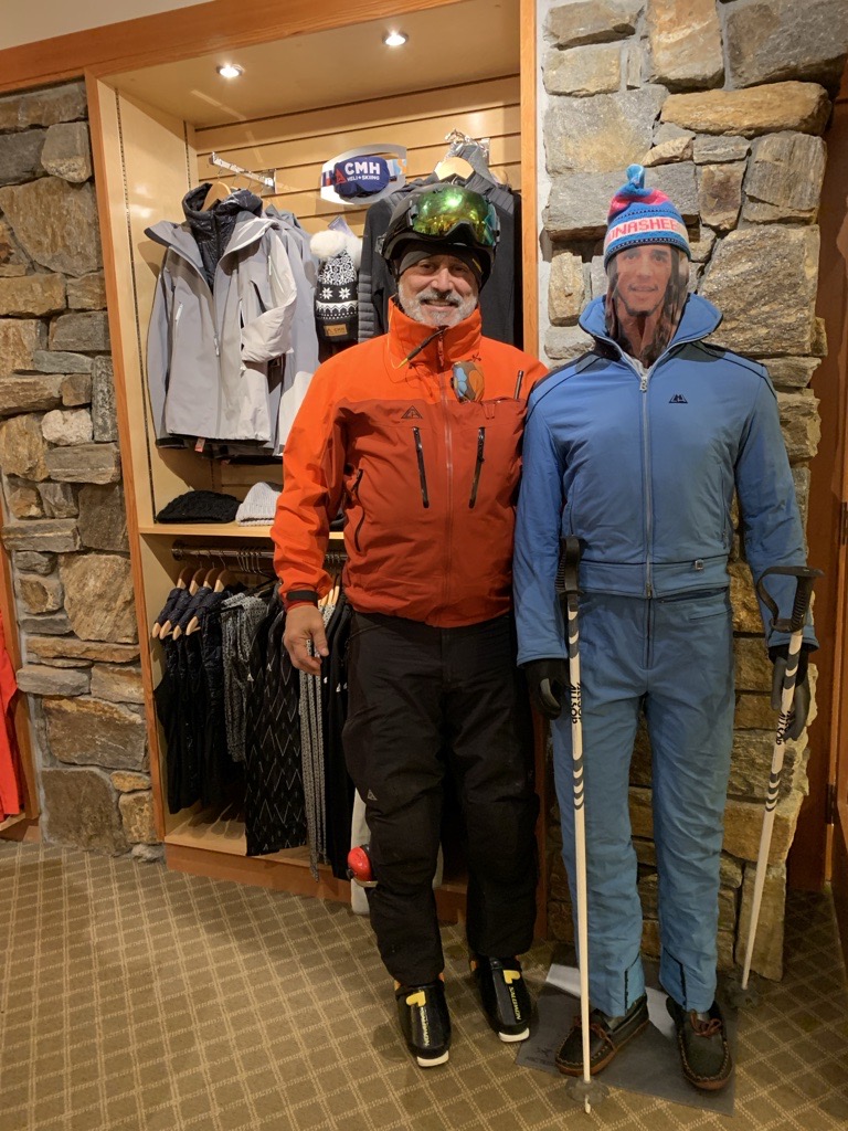 A middle-aged man with a white beard stands in the lobby of a CMH Heli-Skiing lodge, posing next to a mannequin. The mannequin is wearing a vintage blue ski suit. The man is wearing an orange ski jacket, black ski pants, a ski helmet, and goggles. 