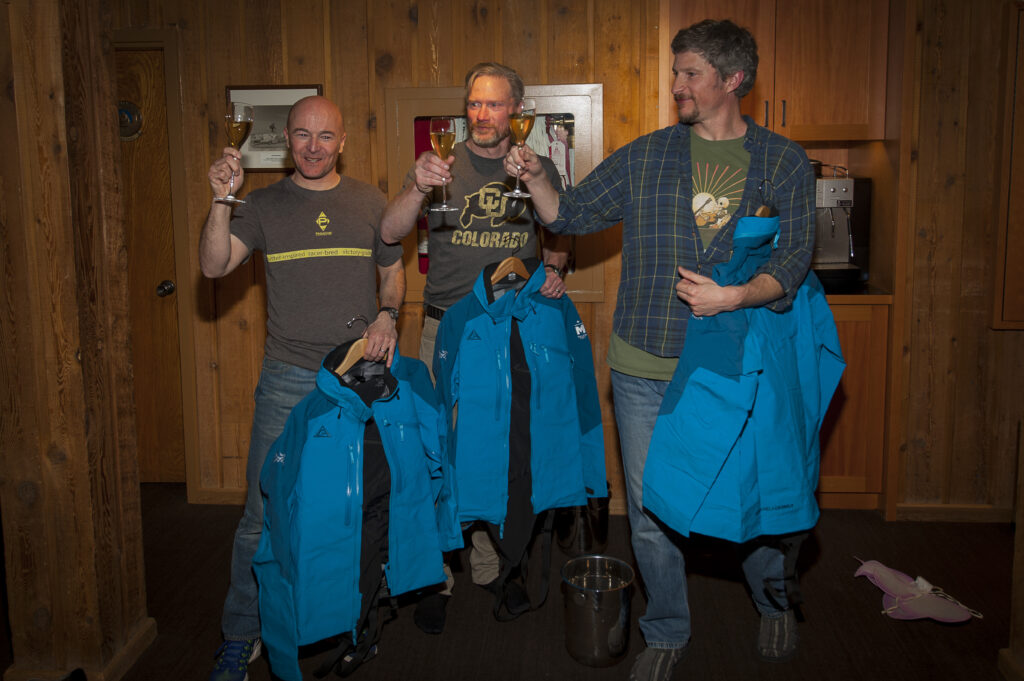 Three middle-aged men dressed in jeans and casual shirts stand next to each other in the dining room of a CMH Heli-Skiing lodge. They are each holding a glass of champagne in one hand and a new blue Million Foot jacket in the other. 