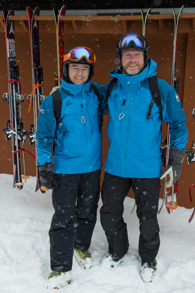 Two male heli-skiers pose for a photo in front of a ski rack at a CMH Heli-Skiing lodge. It's snowing outside and the men are both wearing blue Million Foot ski jackets made by Arc'teryx.