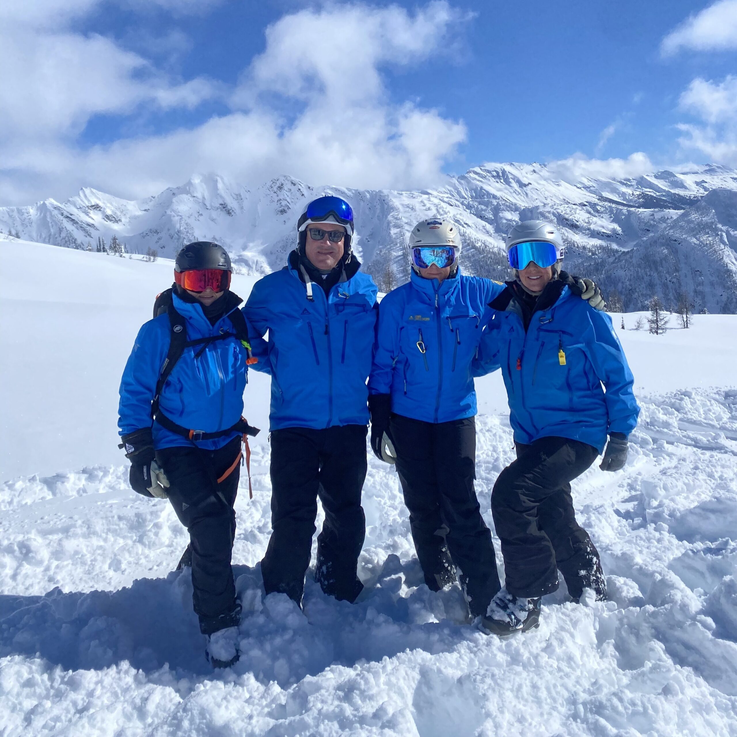 Four friends who heli-ski together pose for a photo while out skiing. They are all wearing matching blue Million Foot jackets from CMH Heli-Skiing. They are standing in powder snow with a mountain range and blue sky behind them. 