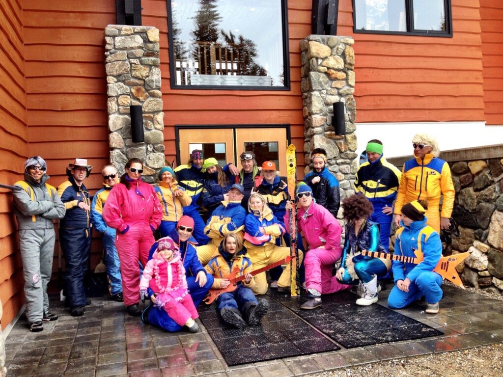 A group of approximately 20 people wearing brightly-coloured, vintage ski suits poses in front of a CMH Heli-Skiing lodge. 