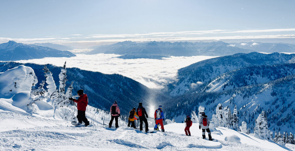 A group of skiers and snowboarders standing on a snowy ridge overlooking a vast mountain range with deep valleys and a sea of clouds below.