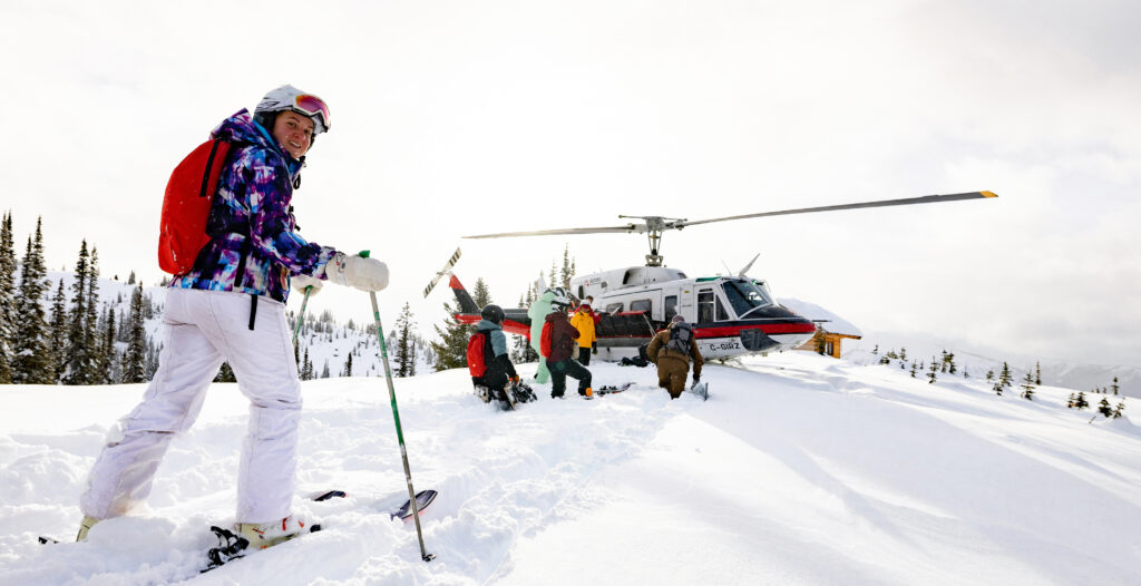 Several skiers and snowboarders walking through deep snow toward a helicopter parked on a snowy ridge.