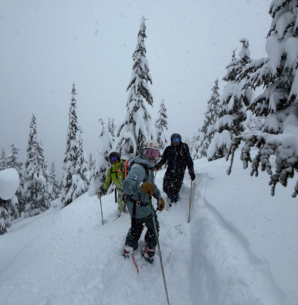 Snow falls over three people standing on a steep, snow‑covered mountainside, surrounded by tall evergreen trees heavily coated in fresh snow.