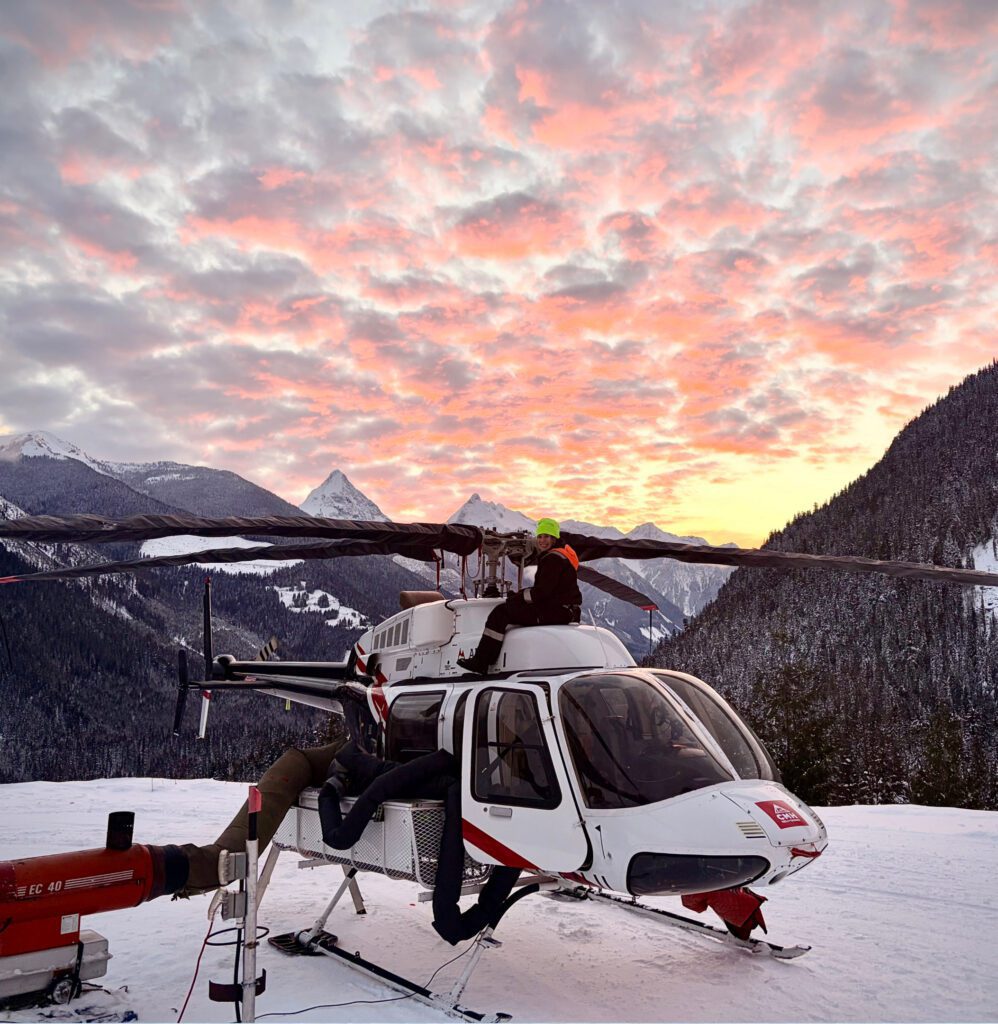 A white helicopter with red markings sits on a snowy mountain plateau at sunset, with one person in a bright green beanie perched on top of the aircraft.