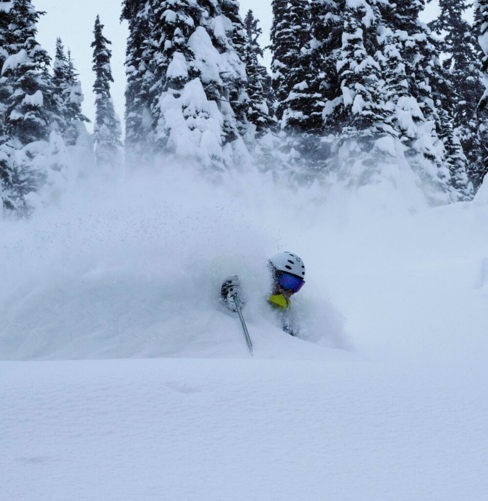 A skier in a bright yellow jacket carves through deep powder, almost completely obscured by a billowing spray of snow.