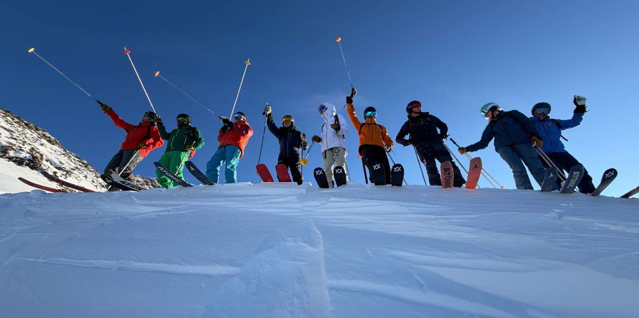 Eight skiers stand in a row on a snowy slope, raising their poles overhead against a bright blue sky.