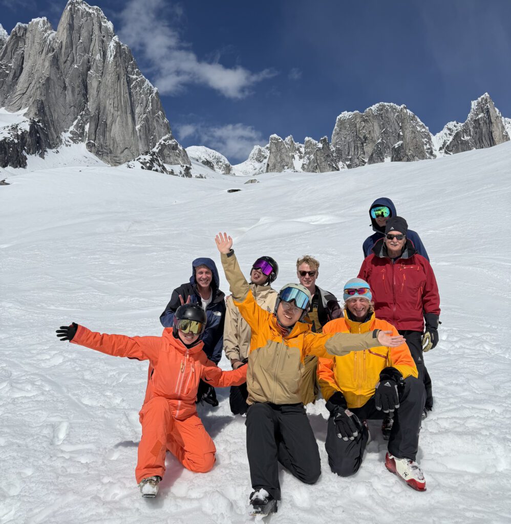 Seven people in colorful ski gear pose together on a snowy mountain slope, with jagged rocky peaks rising behind them under a partly cloudy sky.