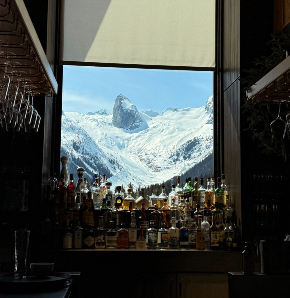 A bar interior window frames a bright mountain view of the towering, snow‑covered Bugaboo spires.