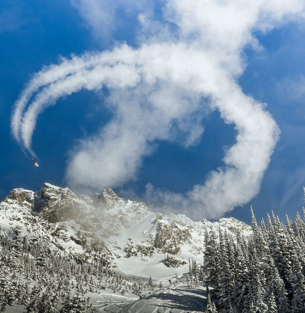 A helicopter draws a perfect white smoke loop high above a snowy mountain landscape