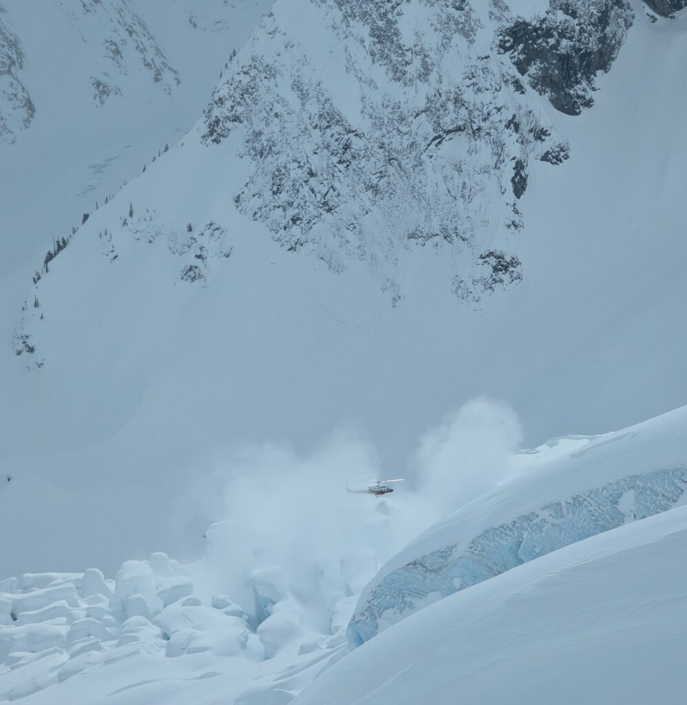A helicopter hovers above a steep, snow‑covered mountainside, kicking up a swirling cloud of powder.