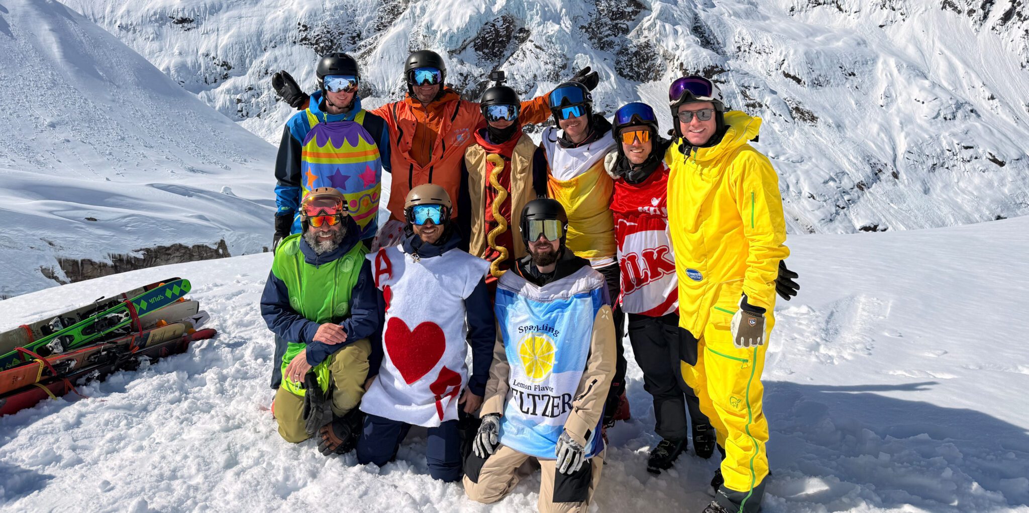 A group of people stand on a snowy mountain slope, dressed in colorful ski and snowboard gear, wearing playful costumes—including a hot dog, a milk carton, a lemon‑seltzer label, and a heart‑themed card