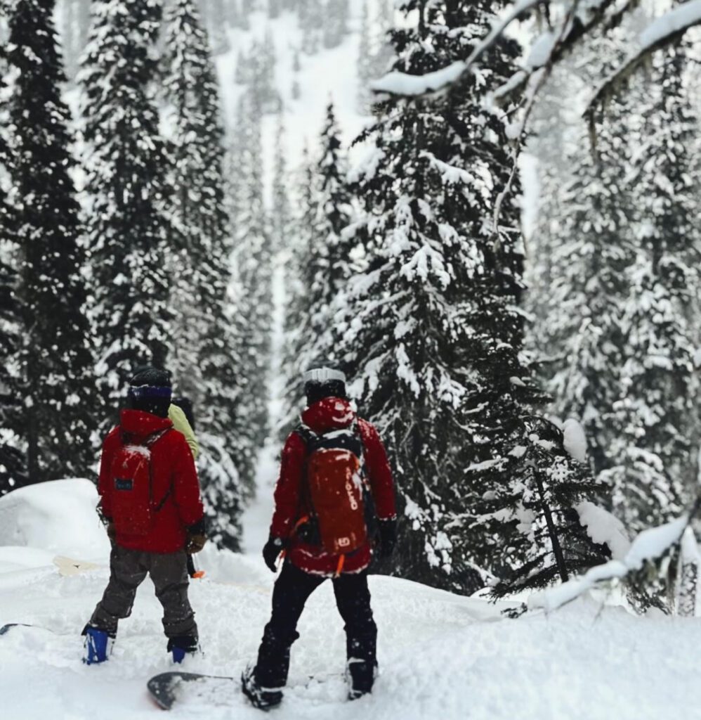 Two snowboarders in red jackets stand on a snowy slope surrounded by tall evergreens coated in fresh snow, their boards resting upright beside them as they pause in a quiet, forested winter landscape.