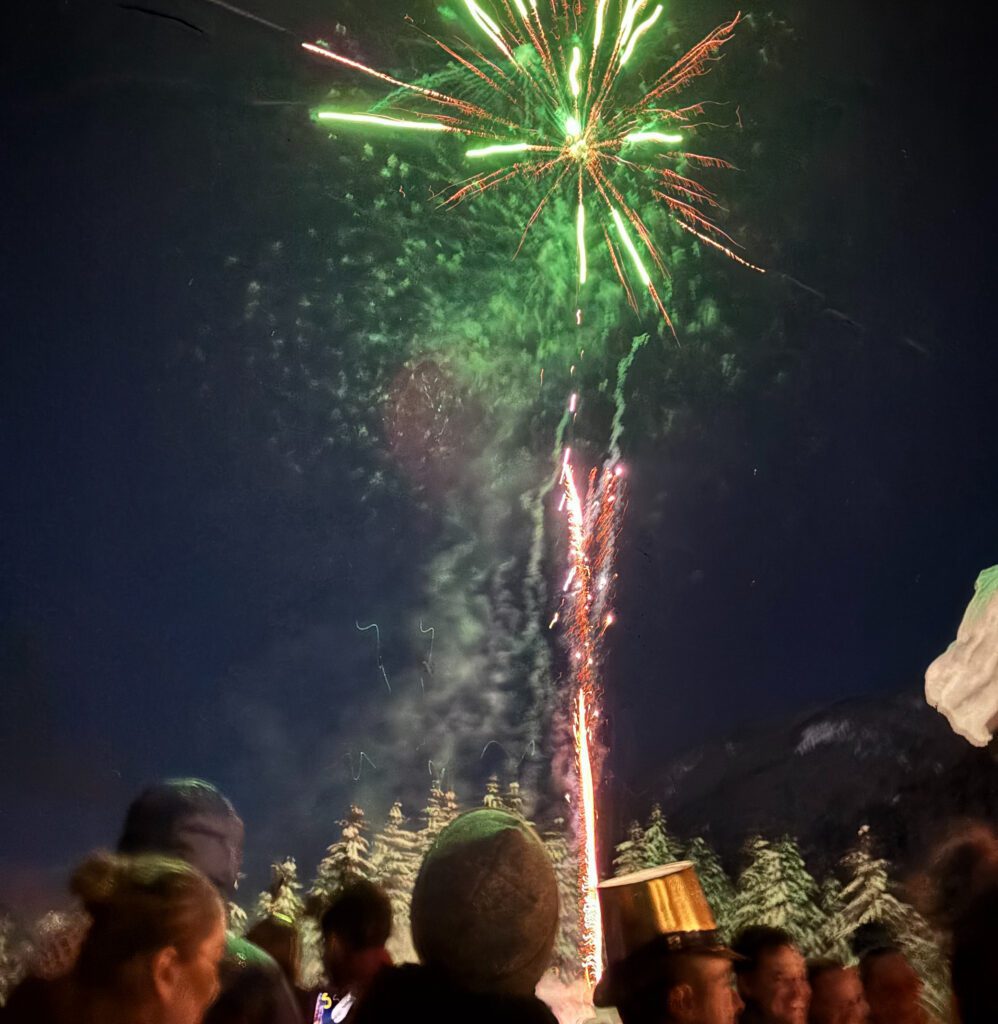 A crowd bundled in winter clothing watches bright green and red fireworks burst overhead