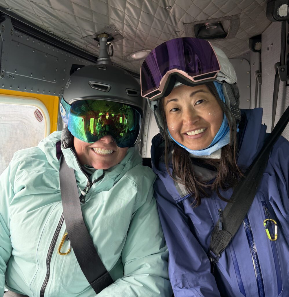 Two people sit inside a helicopter wearing ski and snowboard gear, each buckled into a seat with helmets, goggles, and winter jackets.