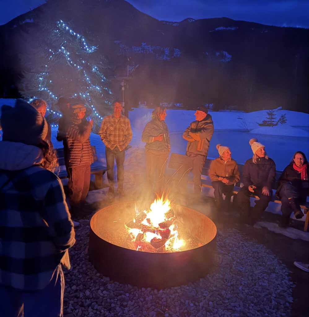 A group of people gather around a large circular fire pit glowing with burning logs, bundled in winter jackets and hats as snow covers the ground.