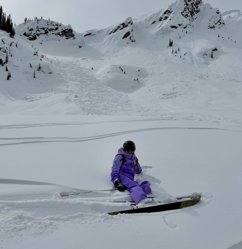 A skier in a purple outfit sits in deep snow on a mountain slope, skis still attached, with winding tracks leading up toward rugged, snow‑covered peaks under an overcast sky.