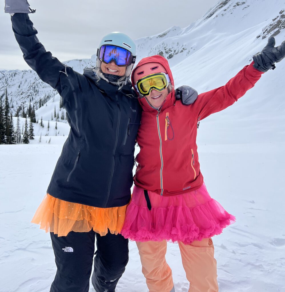 Two people in winter gear stand on a snowy mountain slope, each wearing a colorful tutu—one orange, one bright pink, raising their arms joyfully.