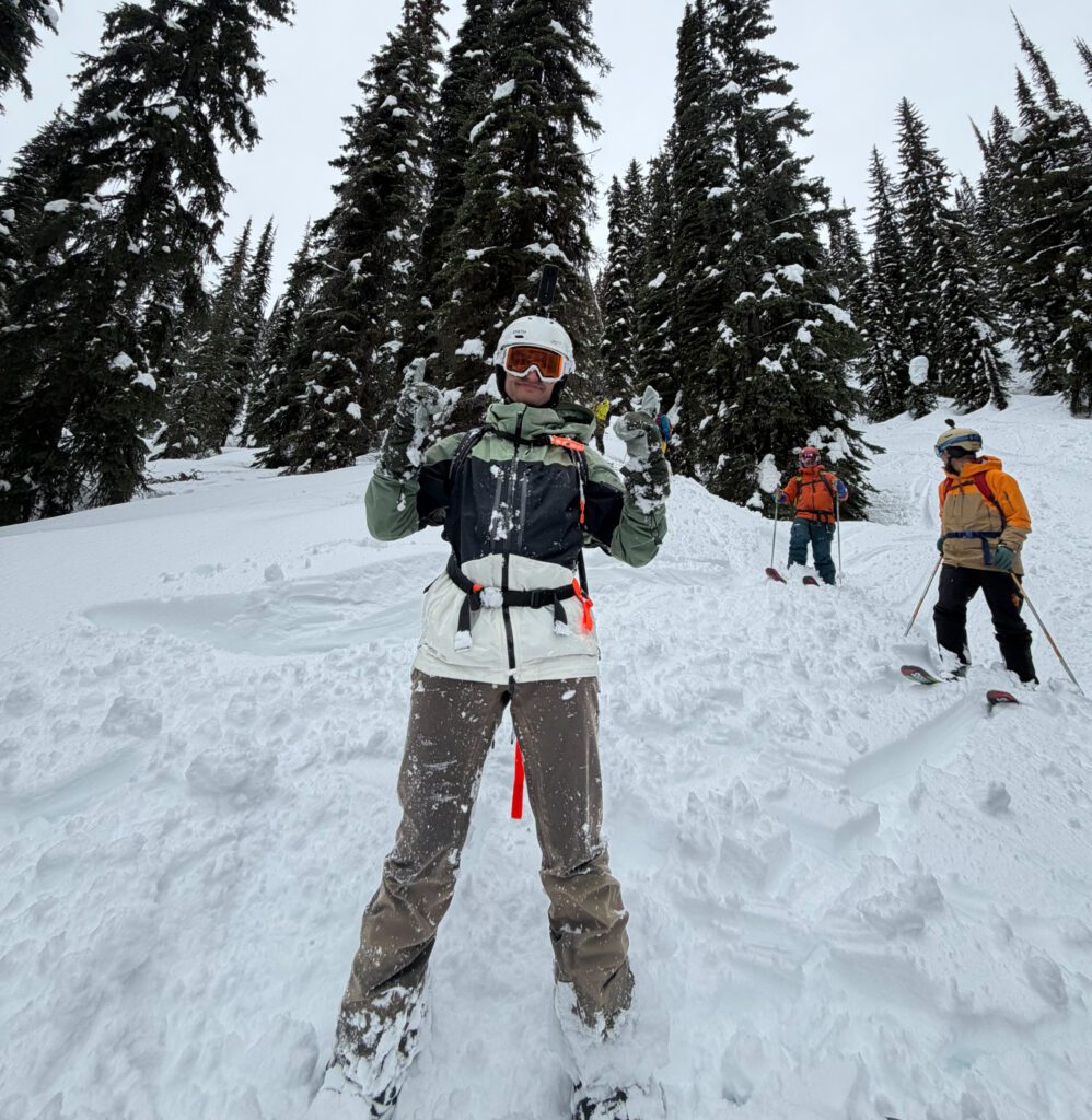 Three skiers stand on a snowy slope among tall, snow‑covered evergreens. The person closest to the camera faces forward with both hands raised