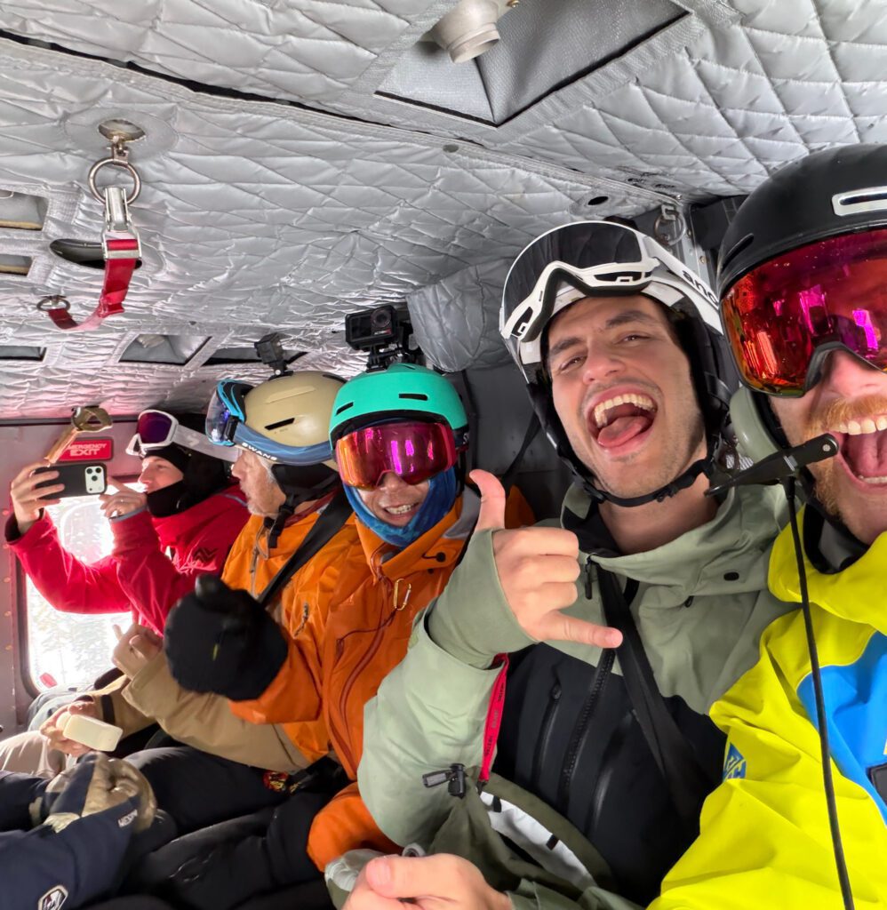 A group of people in ski and snowboard gear sit inside a helicopter, smiling and posing.