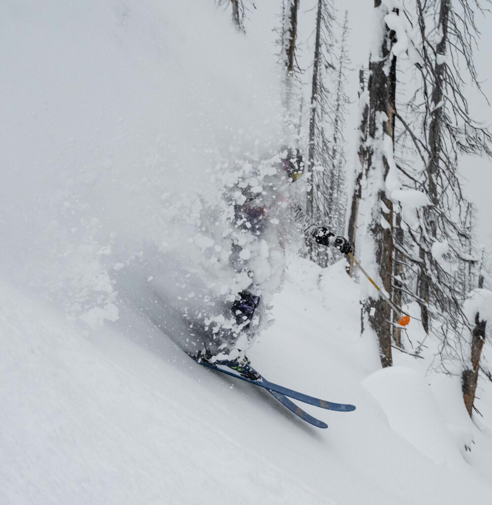 A skier drops down a steep slope in deep powder, almost completely hidden behind a burst of snow.