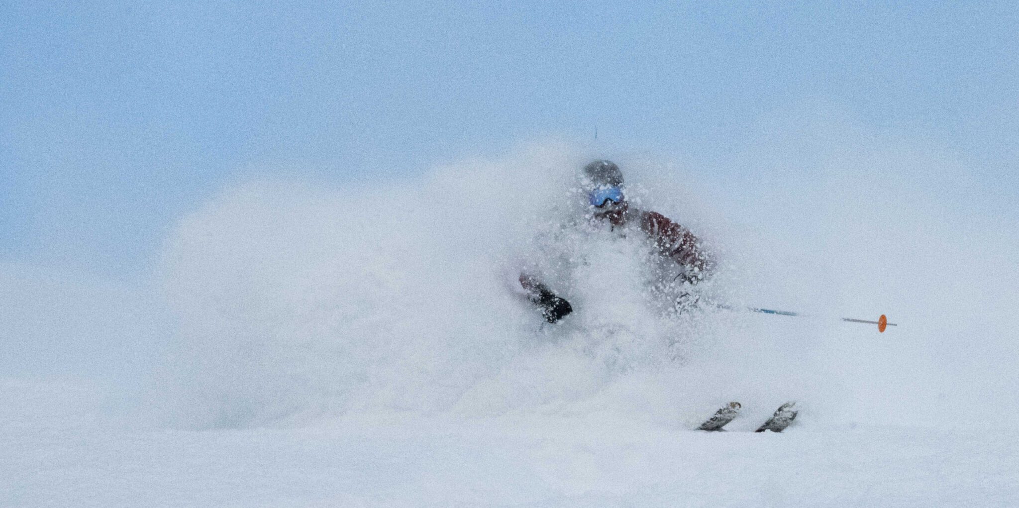 A skier charges through deep powder, almost completely hidden behind a burst of snow, with only their helmet, goggles, and the tips of their poles visible against a pale blue sky.