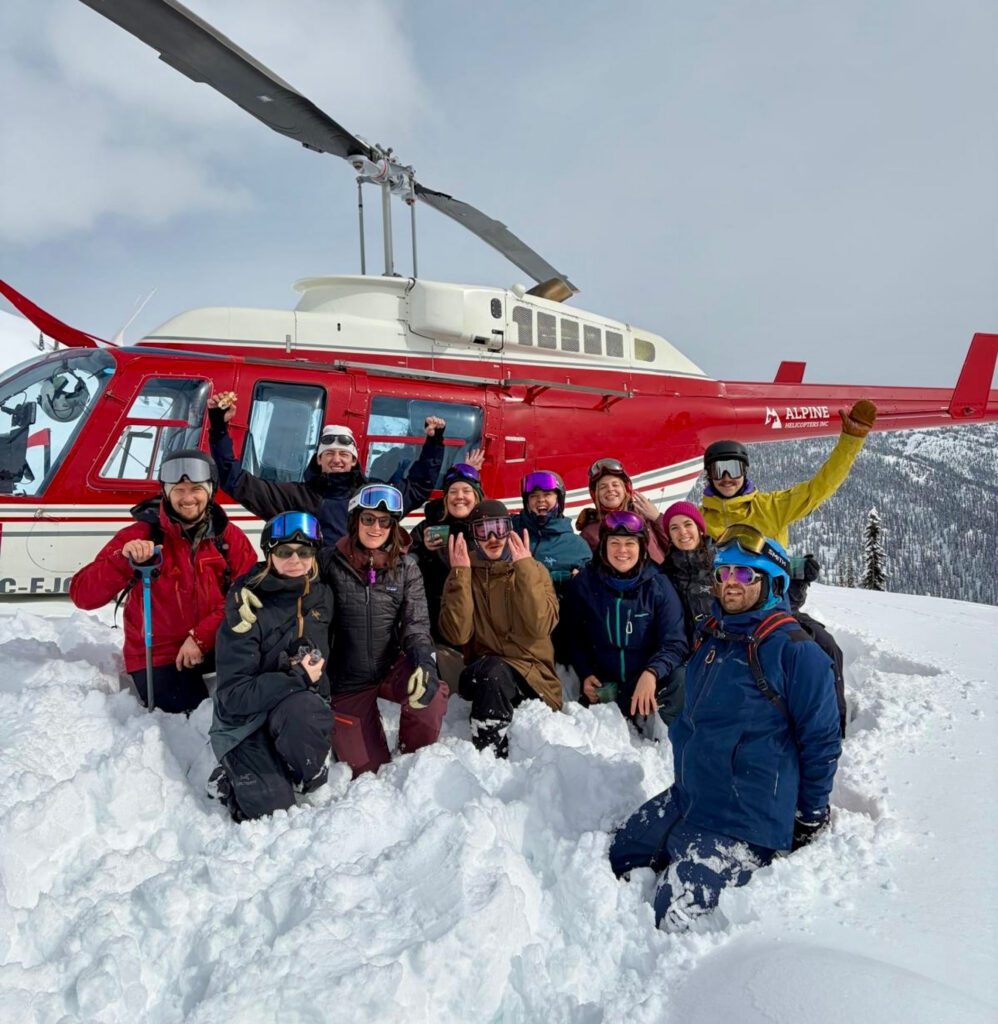 A group of people in winter gear kneel and stand in deep snow in front of a red helicopter, with snowy mountains and trees rising behind them under a cloudy sky.